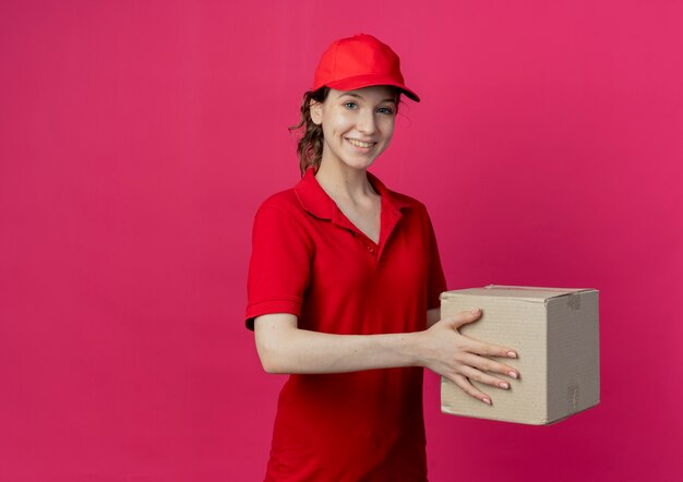 Glimlachend jong mooi leveringsmeisje in rood uniform en de kartondoos van de GLBholding die op karmozijnrode achtergrond met exemplaarruimte wordt geïsoleerd
