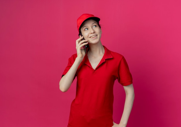 Glimlachend jong mooi leveringsmeisje in rood uniform en GLB die op telefoon spreken die kant bekijken die op karmozijnrode achtergrond met exemplaarruimte wordt geïsoleerd