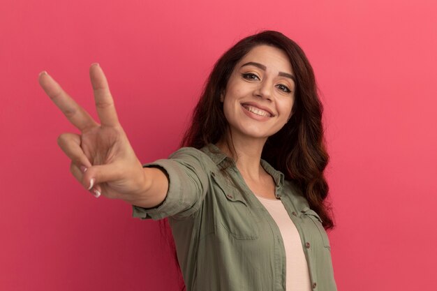 Glimlachend jong mooi meisje dat olijfgroene t-shirt draagt die vredesgebaar toont dat op roze muur wordt geïsoleerd