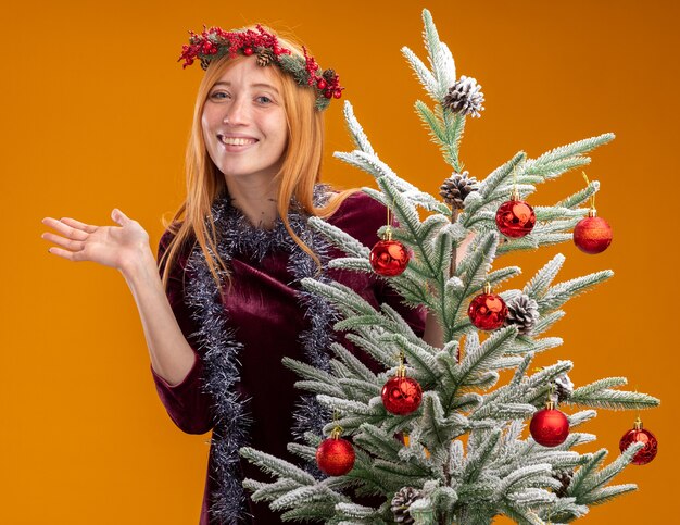 Glimlachend jong mooi meisje dat zich achter de kerstboom bevindt die rode kleding en krans met slinger op hals spreidt die hand op oranje achtergrond wordt geïsoleerd