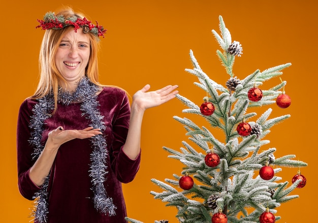 Glimlachend jong mooi meisje dat zich dichtbij kerstboom bevindt die rode kleding en krans met slinger op hals draagt wijst op boom die op oranje achtergrond wordt geïsoleerd
