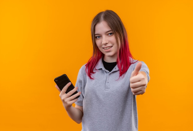 Glimlachend jong mooi meisje die grijze t-shirt dragen die telefoon haar duim op geïsoleerde gele achtergrond houden