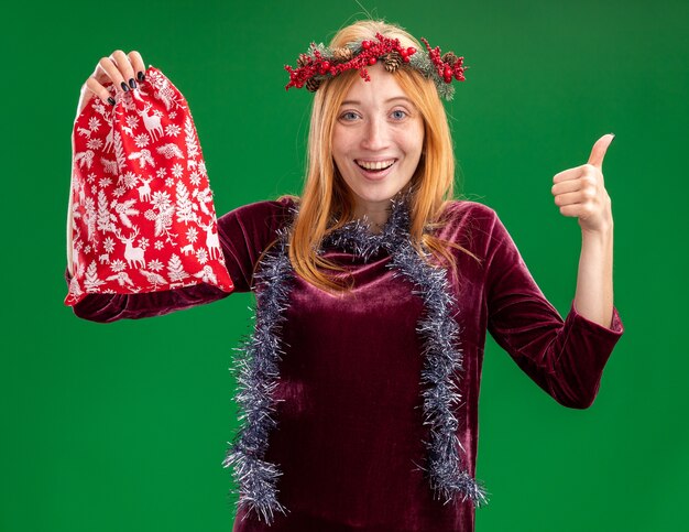 Glimlachend jong mooi meisje die rode jurk met krans en slinger dragen op de nek met kerst tas met duim omhoog geïsoleerd op groene achtergrond