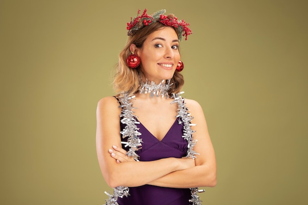 Gratis foto glimlachend jong mooi meisje draagt paarse jurk en krans met guirlande op nek met kerstballen op oren die handen kruisen geïsoleerd op olijfgroene achtergrond