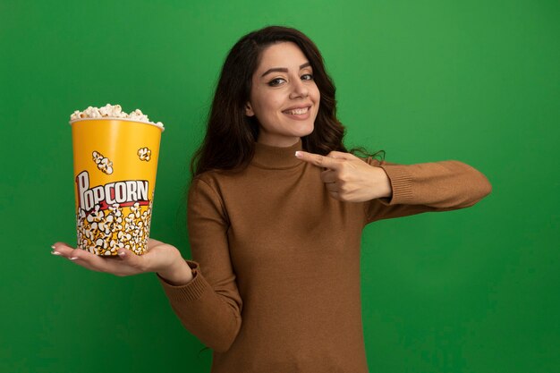 Glimlachend jong mooi meisje houdt en wijst naar emmer popcorn geïsoleerd op groene muur green