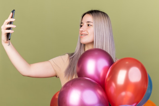 Glimlachend jong mooi meisje met een beugel die een telefoon vasthoudt en kijkt achter ballonnen