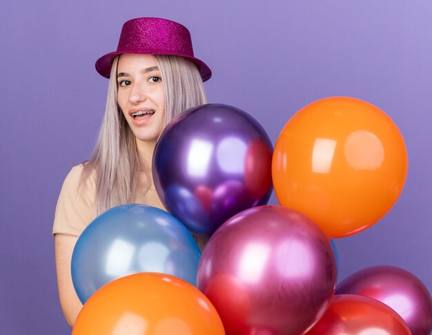 Glimlachend jong mooi meisje met feestmuts die achter ballonnen staat