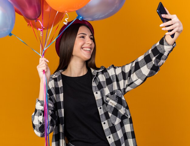 Glimlachend jong mooi meisje met feestmuts die ballonnen vasthoudt en een selfie neemt geïsoleerd op een oranje muur