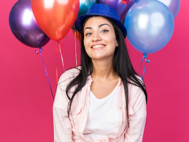 Glimlachend jong mooi meisje met feestmuts die voor ballonnen staat