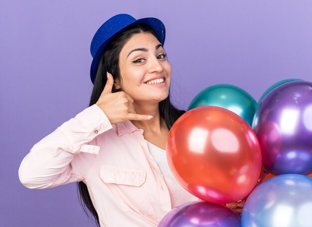 Glimlachend jong mooi meisje met feestmuts met ballonnen die een telefoongesprekgebaar tonen