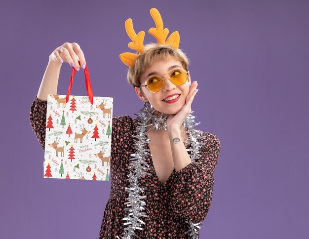 Glimlachend jong mooi meisje met rendiergeweien hoofdband en klatergoudslinger om nek met bril met kerstcadeauzakje met hand op gezicht kijkend naar kant geïsoleerd op paarse muur met kopieerruimte