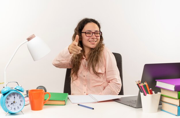 Glimlachend jong mooi schoolmeisje die glazen dragen die aan bureau met schoolhulpmiddelen zitten die haar huiswerk doen die naar voren wijzen en op witte muur knipogen