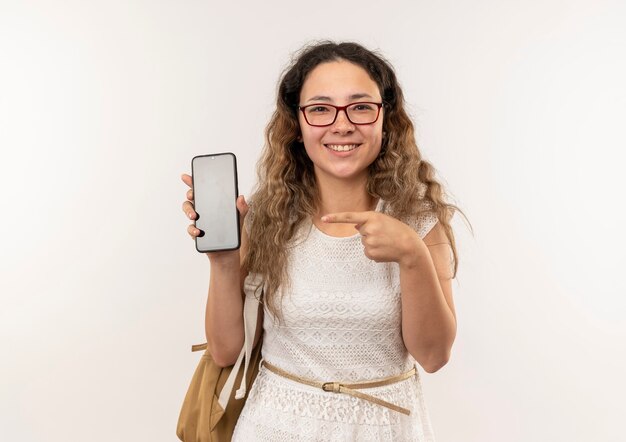 Glimlachend jong mooi schoolmeisje die glazen en achterzak dragen die en op mobiele telefoon tonen richten die op witte muur wordt geïsoleerd