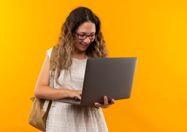 Glimlachend jong mooi schoolmeisje die glazen en achterzak dragen die laptop met behulp van die op gele muur wordt geïsoleerd