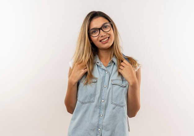 Glimlachend jong mooi studentenmeisje die glazen en de riemen van de achterzakholding van achterzak dragen die op witte muur wordt geïsoleerd