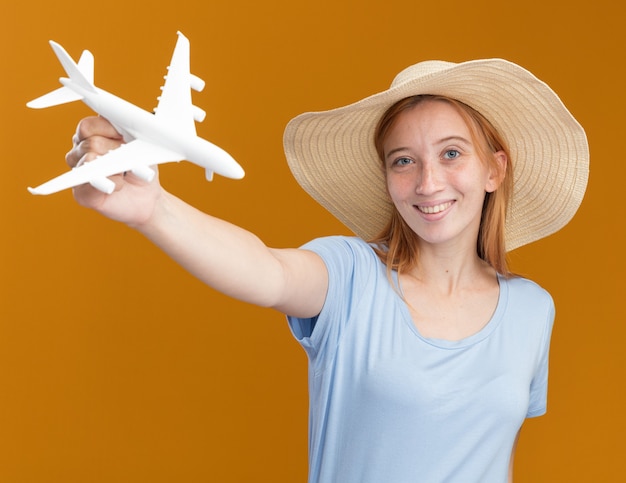 Glimlachend jong roodharig gembermeisje met sproeten met strandhoed houdt modelvliegtuig geïsoleerd op oranje muur met kopieerruimte