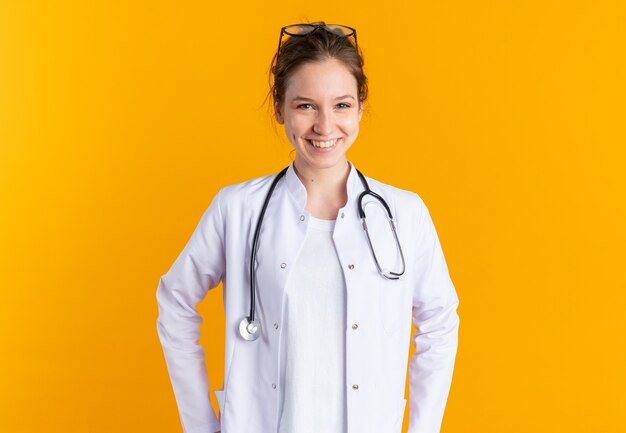 Glimlachend jong Slavisch meisje in doktersuniform met stethoscoop geïsoleerd op oranje muur met kopieerruimte