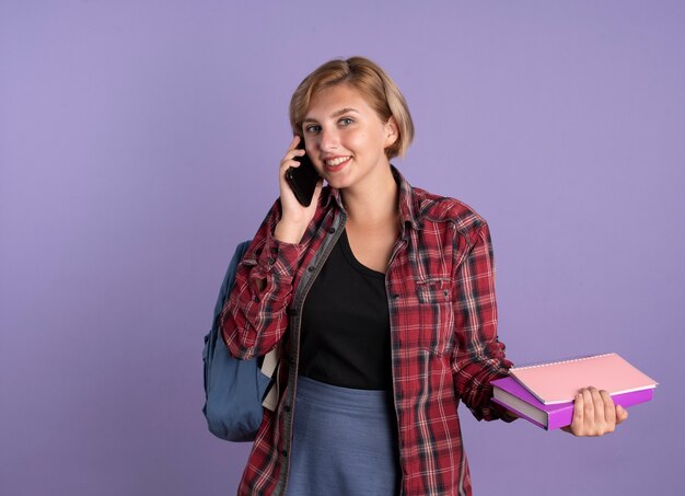 Glimlachend jong slavisch studentenmeisje dat een rugzak draagt, houdt boek- en notitieboekjes aan de telefoon