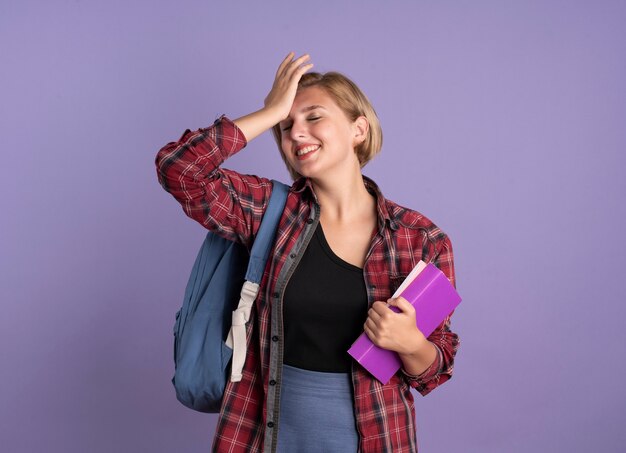 Glimlachend jong slavisch studentenmeisje dat een rugzak draagt, houdt een boek vast en een notitieboekje legt de hand op het voorhoofd
