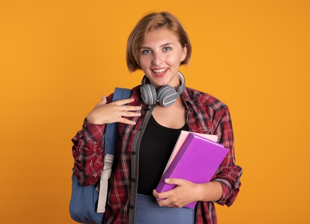 Glimlachend jong slavisch studentenmeisje met hoofdtelefoons die rugzak dragen die boek en notitieboekje houden