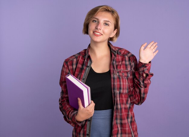 Glimlachend jong slavisch studentenmeisje staat met opgeheven hand houdt boek en notitieboekje vast