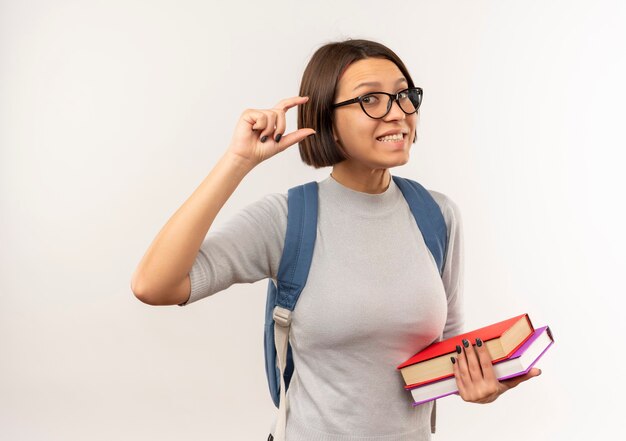 Glimlachend jong studentenmeisje die glazen en de achterboeken van de zakholding dragen die grootte tonen die op witte muur wordt geïsoleerd