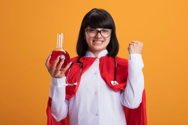 Glimlachend jong superheldmeisje die stethoscoop met medisch gewaad en mantel met glazen dragen die chemieglasfles houden die met rode vloeistof wordt gevuld die ja gebaar tonen