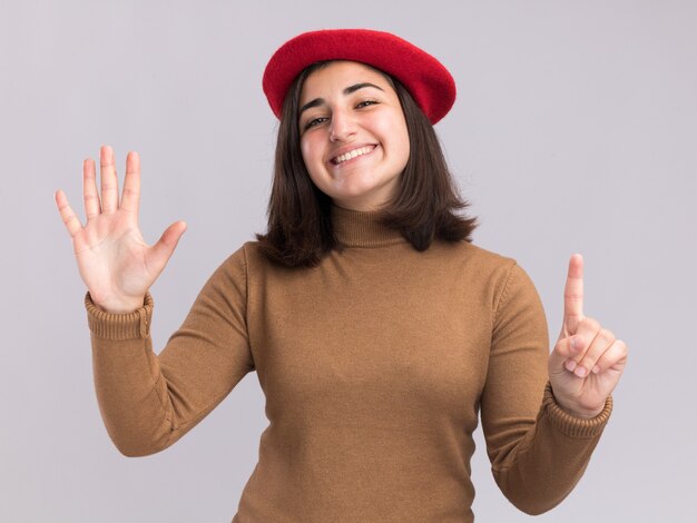 Glimlachend jong vrij Kaukasisch meisje met barethoed die zes met vingers gebaart die op witte muur met exemplaarruimte worden geïsoleerd