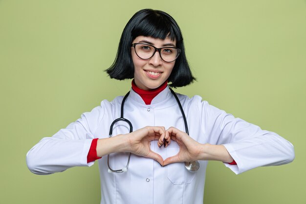Glimlachend jong, vrij kaukasisch meisje met optische bril in doktersuniform met een stethoscoop die een hartteken gebaart