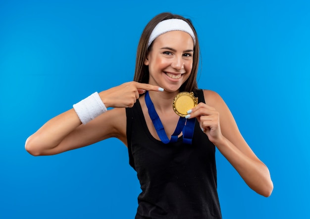 Glimlachend jong vrij sportief meisje dat hoofdband en polsbandje en medaille om haar hals draagt die op medaille richt die op blauwe ruimte wordt geïsoleerd