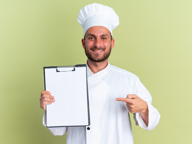 Glimlachend jonge blanke mannelijke kok in uniform van de chef-kok en pet tonen en wijzend op klembord