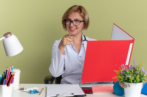 Glimlachend jonge vrouwelijke arts dragen medische gewaad met bril en stethoscoop zit aan tafel met medische hulpmiddelen houden map en wijst op camera geïsoleerd op olijf groene achtergrond