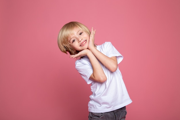 Glimlachend jongensportret van leuk aanbiddelijk blond kind in witte t-shirt en grijze jeans op roze backgorund