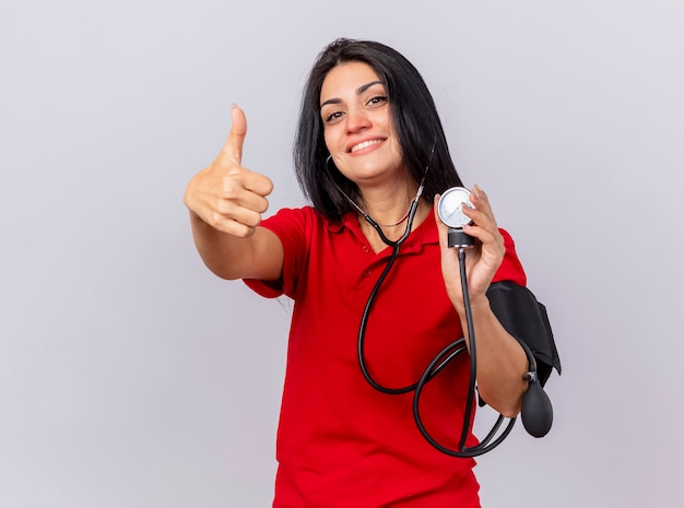 Glimlachend Kaukasisch ziek meisje dragen stethoscoop kijken camera haar druk meten met bloeddrukmeter weergegeven: duim omhoog geïsoleerd op een witte achtergrond met kopie ruimte