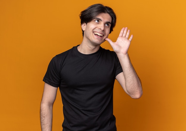 Glimlachend kijkend naar een jonge, knappe kerel met een zwart t-shirt die hand rond het gezicht houdt geïsoleerd op een oranje muur