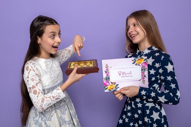 Glimlachend kijkend naar elkaar twee kleine meisjes op een gelukkige vrouwendag met een wenskaart met een doos snoepjes