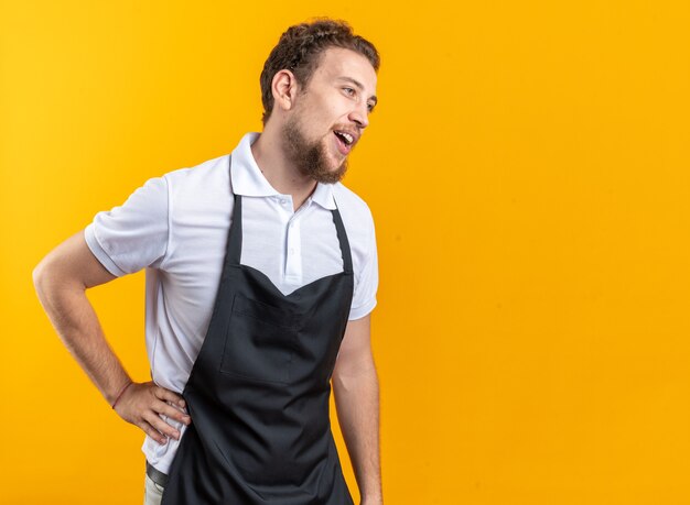 Glimlachend kijkende jonge mannelijke kapper die uniform draagt en hand op de heup legt, geïsoleerd op een gele achtergrond met kopieerruimte