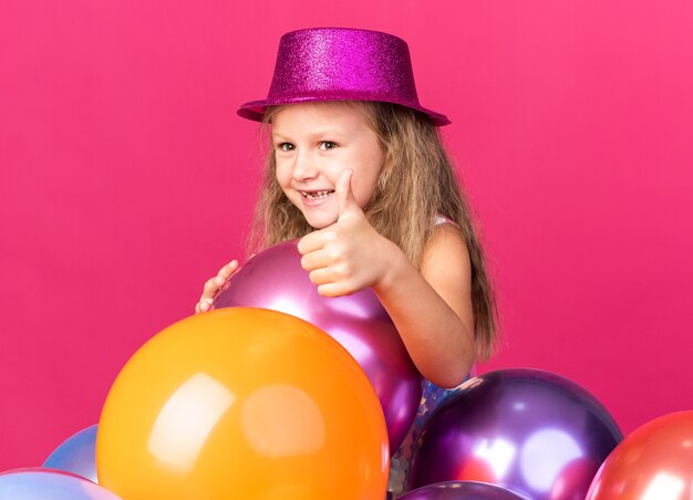 glimlachend klein blond meisje met paarse feestmuts permanent met helium ballonnen thumbing up geïsoleerd op roze muur met kopie ruimte