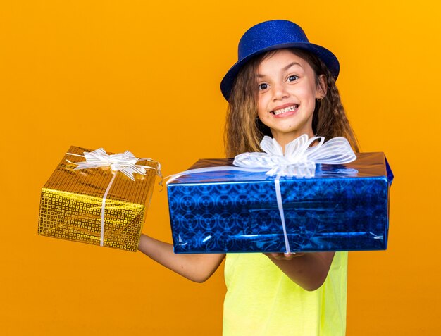 Glimlachend klein kaukasisch meisje met blauwe feestmuts met geschenkdozen geïsoleerd op een oranje muur met kopieerruimte