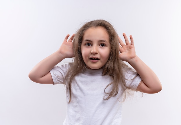 Glimlachend klein schoolmeisje dat een wit t-shirt draagt, legde haar handen om de oren op geïsoleerde witte achtergrond