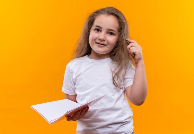 Glimlachend klein schoolmeisje die het witte notitieboekje en de pen van de t-shirtholding op geïsoleerde oranje achtergrond dragen