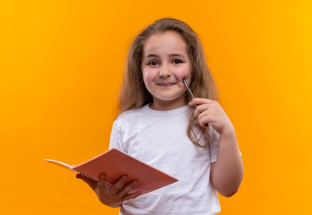 Glimlachend klein schoolmeisje die het witte notitieboekje en de pen van de t-shirtholding op geïsoleerde oranje achtergrond dragen