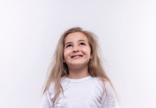 Glimlachend klein schoolmeisje die wit t-shirt dragen die omhoog op geïsoleerde witte achtergrond kijken