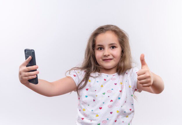 Glimlachend klein schoolmeisje die witte t-shirt dragen die haar duim op geïsoleerde witte achtergrond houden