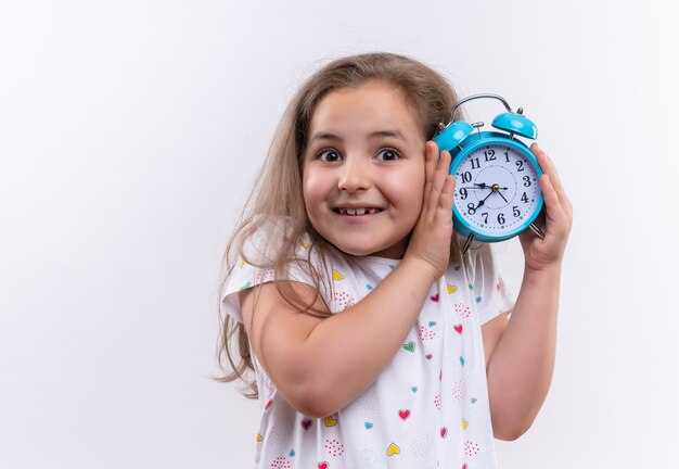 Glimlachend klein schoolmeisje die witte t-shirt dragen die wekker rond oor op geïsoleerde witte achtergrond houden