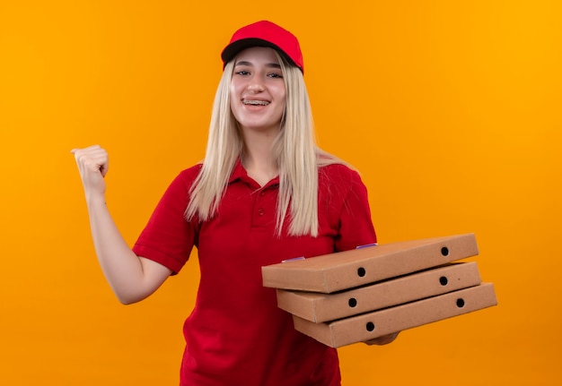 Glimlachend levering jong meisje dat rode t-shirt en pet draagt die pizzadoos toont die ja gebaar op geïsoleerde oranje achtergrond toont