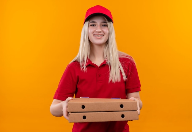 Glimlachend levering jong meisje die rode t-shirt en pet in de pizzadoos van de beugelholding op geïsoleerde oranje achtergrond dragen