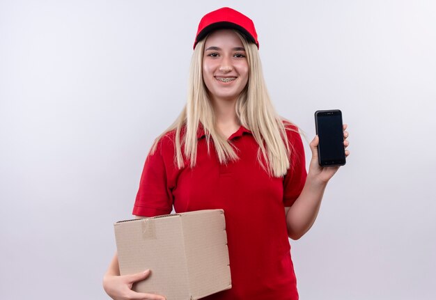 Glimlachend levering jong meisje die rode t-shirt en pet in tandsteun houden telefoon en doos op geïsoleerde witte achtergrond