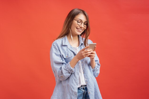 Glimlachend meisje dat in vrijetijdskleding en oortelefoons aan het scherm van de telefoon kijkt