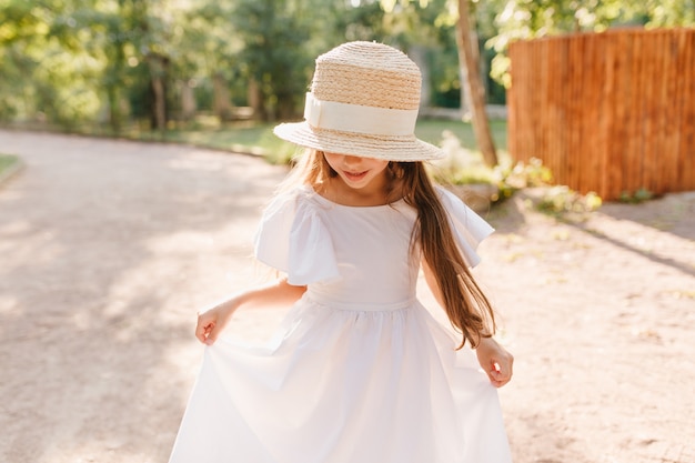 Glimlachend meisje in grote strooien hoed kijkt naar haar voeten tijdens dans in park. Kleine dame draagt een stijlvolle schipper die speelt met een witte jurk en geniet van nieuwe kleding.